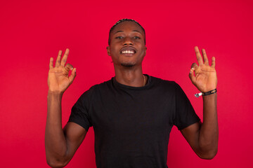 Fototapeta premium Glad Young African American handsome man standing against red background shows ok sign with both hands as expresses approval, has cheerful expression, being optimistic.