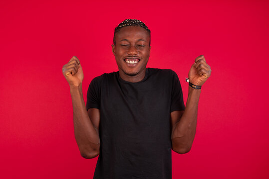 Young African American handsome man standing against red background rejoicing his success and victory clenching fists with joy being happy to achieve aim and goals. Positive emotions, feelings.