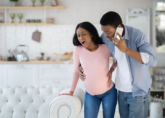 Pregnant African American woman having prenatal contractions, worried husband calling doctor on smartphone - obrazy, fototapety, plakaty