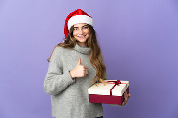 Girl with christmas hat holding a present isolated on white background giving a thumbs up gesture