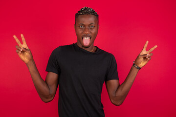 Young African American handsome man standing against red background with optimistic smile, showing peace or victory gesture with both hands, looking friendly. V sign.