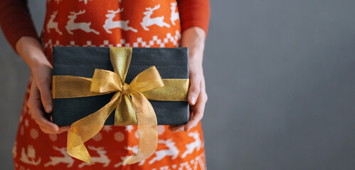 a woman dressed in a red dress and a Christmas apron holds a gift box in front of her.