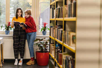Concept of education, library, students and teamwork. Full height of two focused productive stylish high school student girls stands in the library and learning together from notes near sunny window