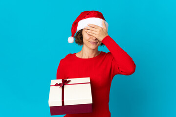 English girl with christmas hat holding a present isolated on blue background covering eyes by hands. Do not want to see something