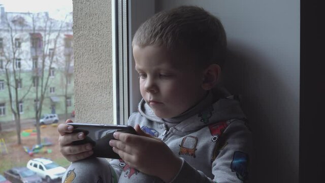 four year old boy with smartphone on windowsill. View of autumn Park and street space for text. Danger of internet