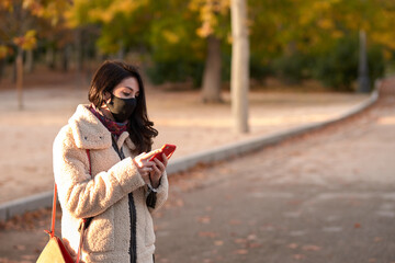 Beautiful woman with facemask sending a text message with her smartphone in a park