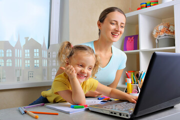 Mother and her daughter sit at the kitchen table studying online together, mom and little girl...