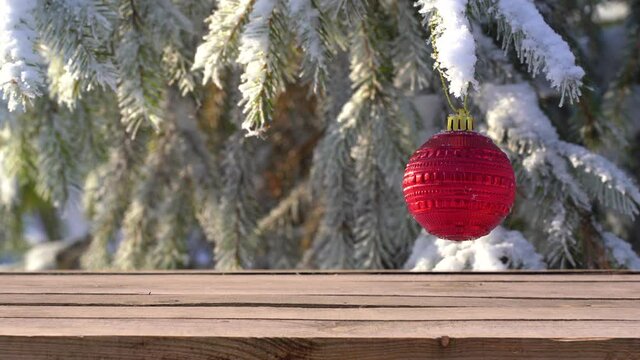 Closeup view video of wooden empty surface of tabel isolated at snowy pine or fir tree branches with one red holdiay ball hanging on branch. Abstract holiday background.