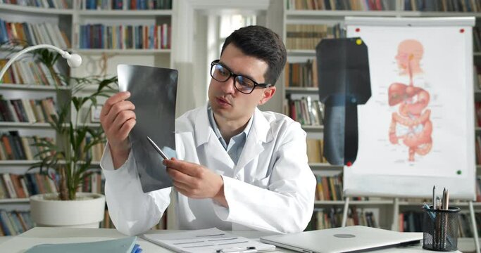 Crop view of man in glasses taking and showing x ray picture to patient while having video call. Male doctor in white professional rob talking and looking to camera in medical office