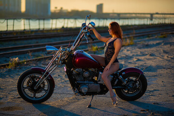 Obraz na płótnie Canvas Red-haired woman in sexy lingerie in high heels sits on a motorcycle. Attractive red-haired girl sits on a motorcycle at sunset