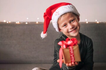 Obraz premium Funny smiling joyful child boy in Santa red hat holding Christmas gift in hand. Christmas concept.