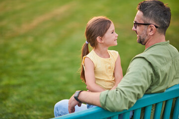 Young father spending time with his cute little daughter. They are looking at each other while sitting on the bench in the park on a summer day