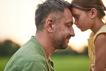 Close up shot of young father and adorable little daughter touching foreheads, standing with eyes closed while spending time outdoors in the park on a summer day