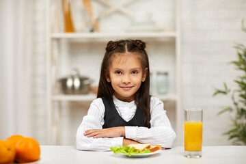 happy little child girl in school uniform having breakfast - sandwich and orange juice in the kitchen. healthy breakfast