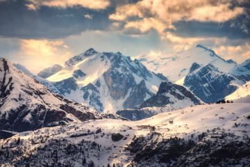 Snowy mountains is lighted by sunbeam at sunset in winter. landscape with beautiful snow covered rocks, buildings on the hill, sky with orange sunlight and clouds. Alps in Dolomites, Italy. Nature
