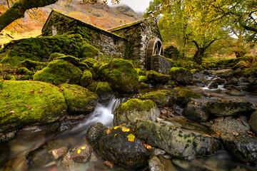 Beautiful Autumn colours at the old mill with waterwheel in Borrowdale, Keswick, Lake District, UK. A small waterfall can be seen in the foreground with moss covered rocks.