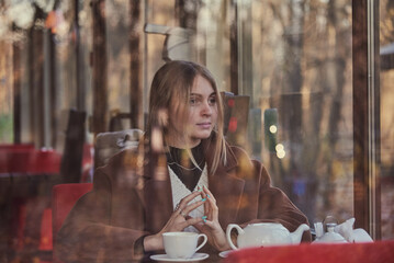 Slightly blurry portrait of a charming girl, made through the window of a cafe. The window reflects the autumn Park.