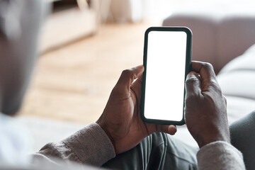 African american man holding smart phone with mockup white blank display, empty screen for app ads sitting on couch at home. Mobile applications technology concept, over shoulder close up view. - Powered by Adobe