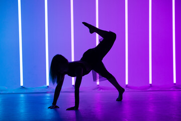Silhouette of a young woman dancer dancing and posing in a room with neon lamps. A slender modern model in a beautiful pose on a purple blue background.
