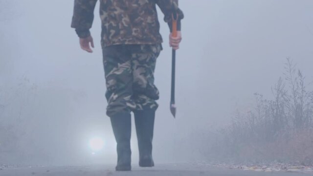 Back view: The maniac&rsquo;s legs are walking along the road with an ax, the headlights of a car