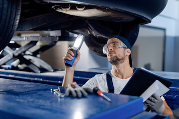 Auto mechanic examining chassis of a vehicle with a flashlight in a workshop. - obrazy, fototapety, plakaty