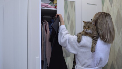 A woman with a cat in her arms opens a wardrobe and chooses what to wear