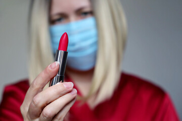 Woman in disposable medical mask looking on red lipstick in her hand. Makeup and cosmetics during the coronavirus pandemic