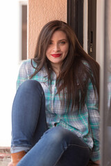 Obraz premium attractive young latin woman with long hair and makeup, happy and sitting on the edge of the window, wearing a plaid t-shirt and jeans