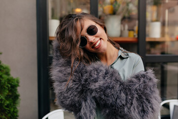 Excited charming woman with wavy hair dressed fur coat in glasses posing at camera