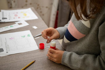 A schoolgirl does her homework. The girl puts glue on the piece of paper to stick to the card. Hands of the girl on the foreground