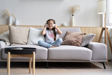 Fototapeta na wymiar Adorable small schoolgirl sitting on sofa, wearing modern wireless headphones, listening to favorite music, Cute little child resting on couch