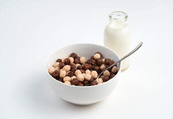 Cereal and chocolate balls in a white bowl and milk in a bottle on a light background. Side view. The concept of healthy, dietary breakfasts.