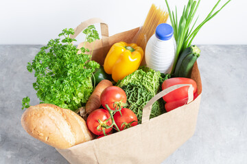 Paper shopping box full of fresh organic vegetables and grocery products on grey table.