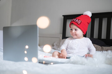 Infant baby in Christmas hat chatting via lap top at home during quarantine. Social distancing during lock down. New year and Christmas celebration at home