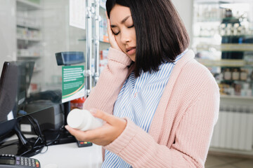 Asian woman suffering from headache and holding bottle with pills in drugstore