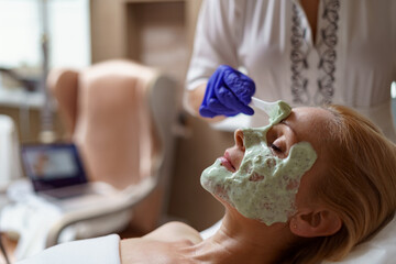 Hands of cosmetology specialist applying facial alginate mask using wooden spatula, making skin...