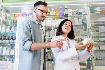 Obraz na płótnie Canvas bearded man pointing with finger at bottles with medication in hands of asian pharmacist in drugstore