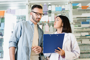 asian pharmacist in white coat holding clipboard near bearded customer in drugstore