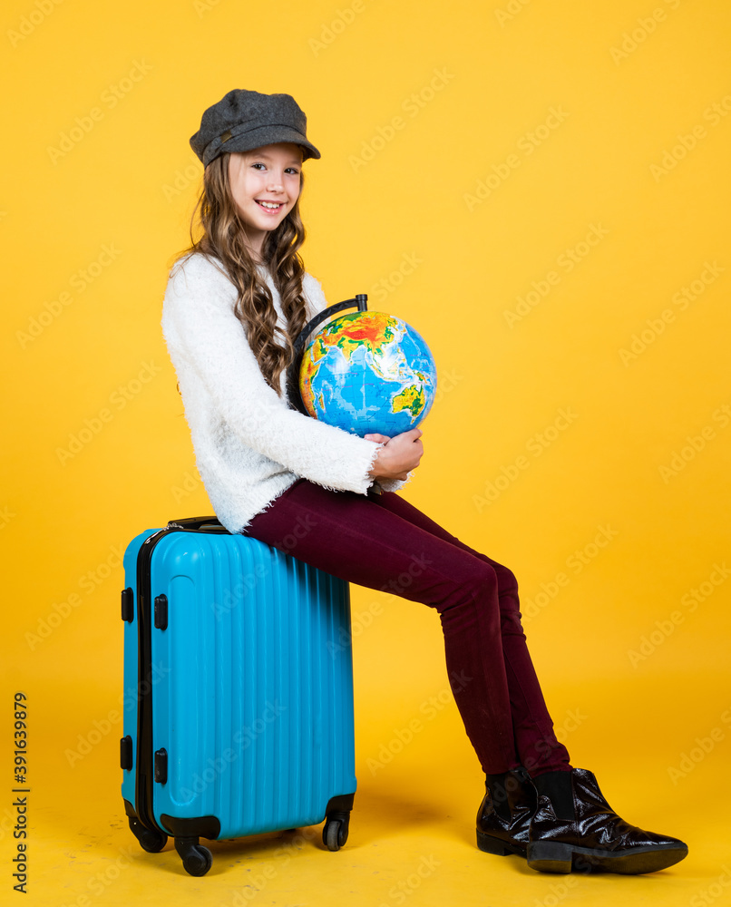 Wall mural cheerful child in headwear with valise for travelling on yellow background, holiday