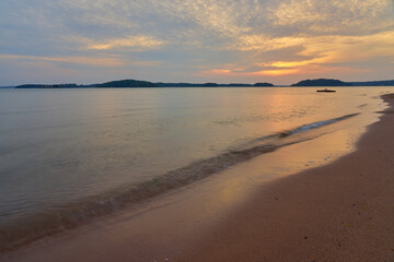 Sunset on the sandy beach with soft waves hitting sand and cloudy sky on the background.