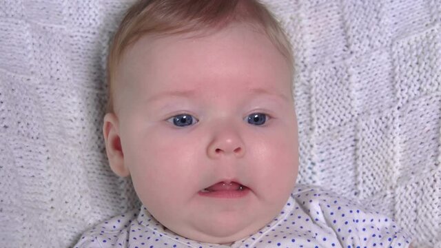 Close-up of a little blue-eyed baby on a white knitted blanket looking at the camera 