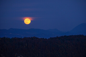 huge orange full moon rising behind a mountain ridge illuminating misty clouds creating a mystical...
