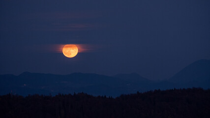 huge orange full moon rising behind a mountain ridge illuminating misty clouds creating a mystical...