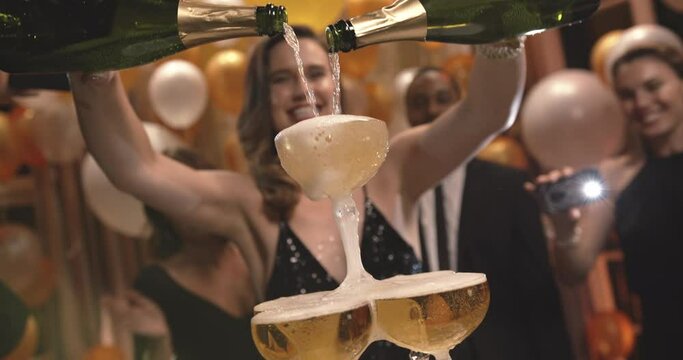 Excited young Woman filling champagne into a tower glasses with friends dancing around and shooting a film on their mobile phone at gala night party. Group of people enjoying at new years party.
