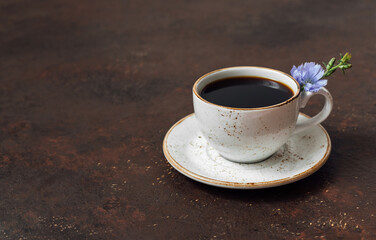 Chicory drink in a cup and a blue chicory flower on a dark brown background. Coffee substitute. Space for text.