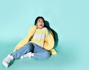 Happy laughing asian kid girl in stylish yellow furry coat, jeans and sneakers sits sprawled on the floor and looks up