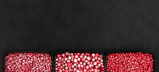 Border of frozen berries in containers on a black background. Cranberries, lingonberries, red currants, berry mix. Copy space, view from above.