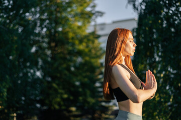 Side view of attractive redhead young woman with closed eyes practicing yoga and meditation performing namaste pose. Calm female making Namaste gesture outside in city park in evening at sunset.