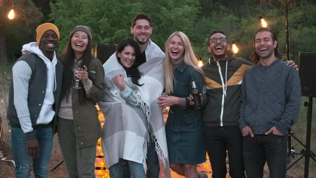 Medium close up of multi-ethnic best friends standing in row with drinks having open air summer party on cool evening laughing and having fun together looking at camera