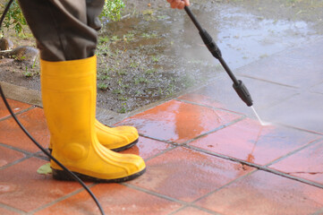 Person wearing yellow rubber boots with high-pressure water nozzle cleaning the dirt in the tiles